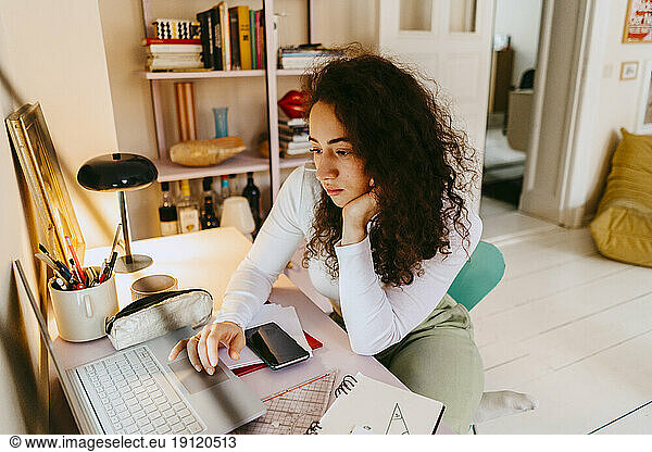 Junge Frau mit lockigem Haar  die einen Laptop benutzt  während sie zu Hause lernt