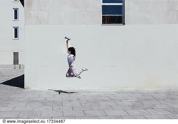 Junge Frau mit Megaphon springt an einem sonnigen Tag an einem Gebäude vorbei