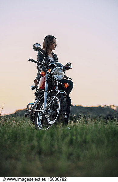 Junge Frau mit Oldtimer-Motorrad in ländlicher Szene genießt Sonnenuntergang