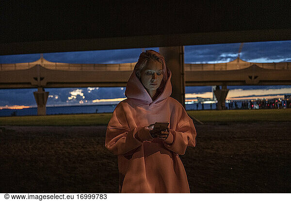 Junge Frau mit rosa Haaren und rosa Kapuzenshirt hält Smartphone in der Stadt bei Nacht