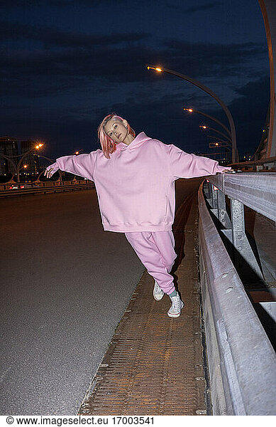Junge Frau mit rosa Haaren und rosa Trainingsanzug steht nachts an einer Straßenschranke