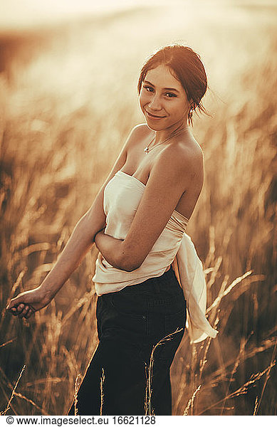 Junge Frau mit schulterfreiem Oberteil steht in einem Feld bei Sonnenuntergang