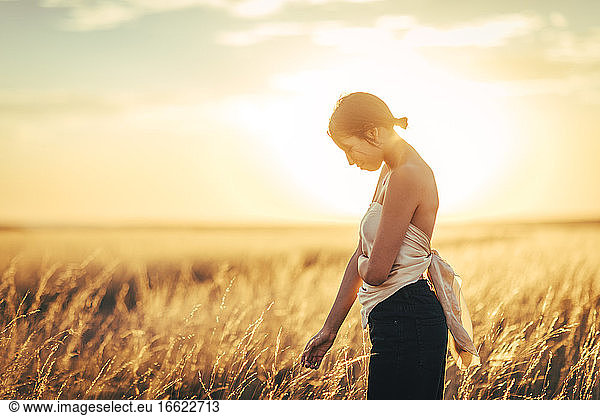 Junge Frau mit schulterfreiem Oberteil steht in einem Feld gegen den Himmel bei Sonnenuntergang
