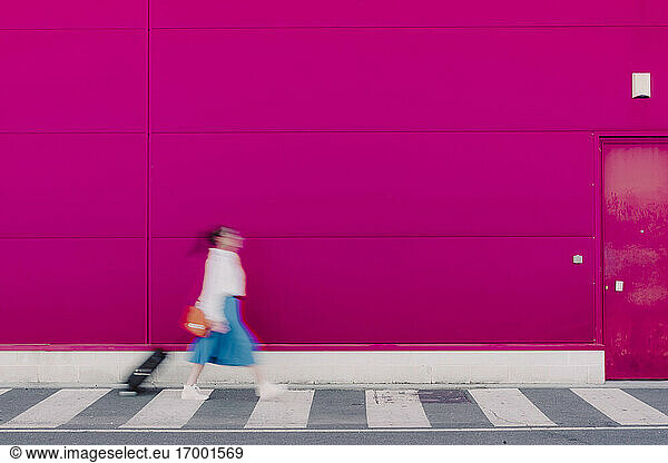Junge Frau mit Smartphone geht mit Trolley entlang einer rosa Wand  unscharf