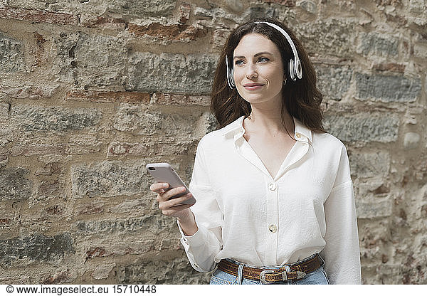 Junge Frau mit Smartphone und Kopfhörern im Freien an einer Steinmauer