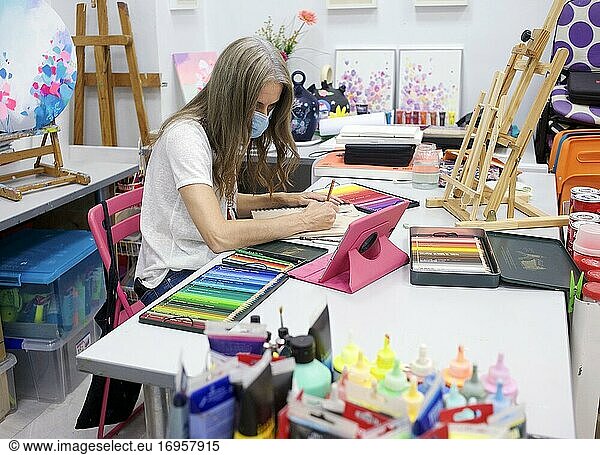 Junge Frau mit weißem Hemd sitzt im Atelier und arbeitet an einem Bild.