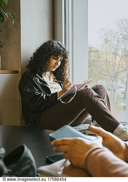 Junge Frau schreibt in ein Buch  während sie in der Universität am Fenster sitzt