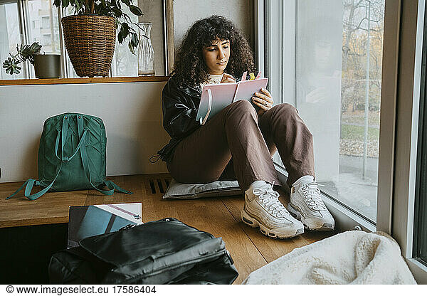 Junge Frau schreibt in voller Länge in ein Buch  während sie in der Universität am Fenster sitzt