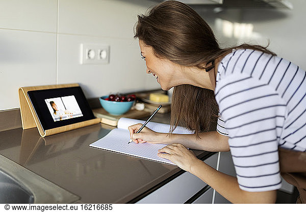 Junge Frau sieht sich ein Video über ein digitales Tablet an und schreibt zu Hause ein Rezept in ein Buch