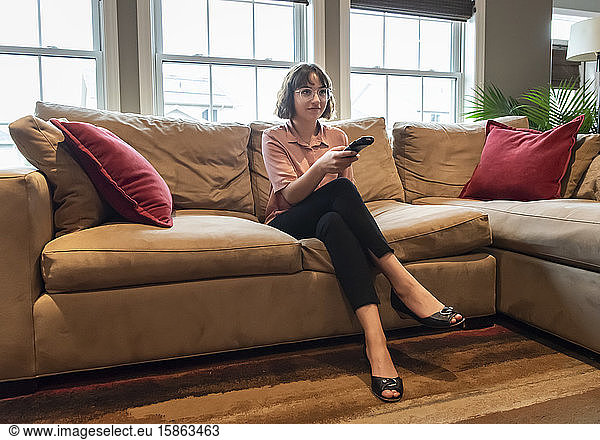 Junge Frau sitzt in Geschäftskleidung auf einer Couch und sieht fern.