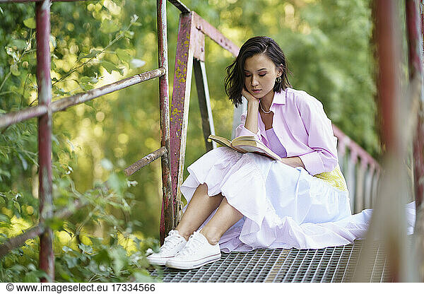 Junge Frau sitzt mit der Hand am Kinn und liest ein Buch auf einer Fußgängerbrücke auf dem Lande
