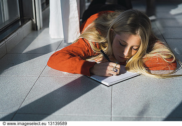 Junge Frau skizziert auf Buch  während sie zu Hause auf dem Fliesenboden liegt