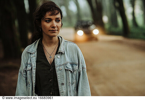 Junge Frau steht allein auf einem Waldweg mit einem Auto dahinter
