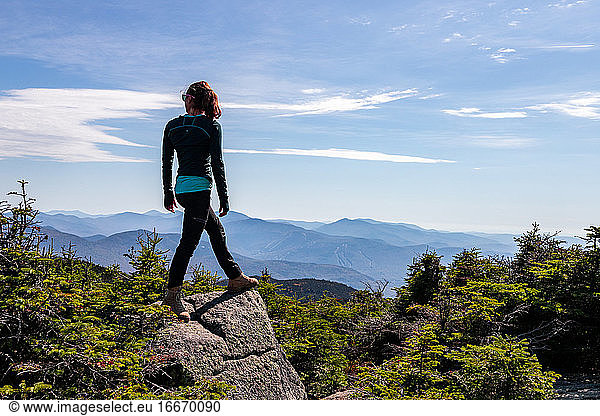 Junge Frau steht auf dem Gipfel eines Berges und blickt auf die dahinter liegenden Gipfel.