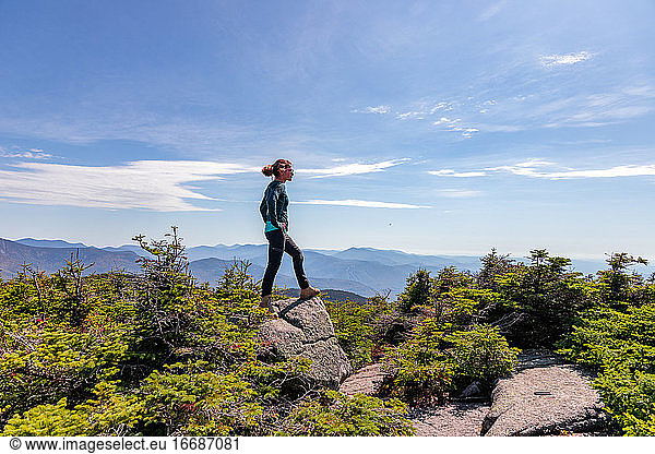 Junge Frau steht auf dem Gipfel eines Berges und blickt auf die dahinter liegenden Gipfel.