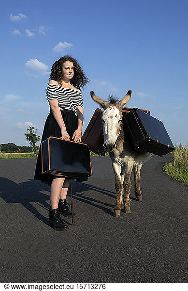 Junge Frau steht auf Landstraße mit Esel  der Gepäck trägt