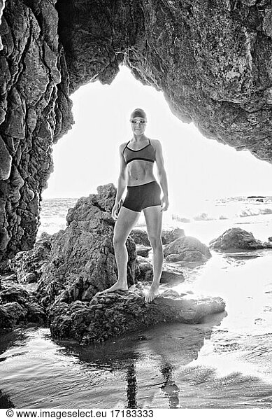 Junge Frau  Triathletin im Training in Badekappe und Schwimmbrille durch einen Felsbogen am Strand gesehen