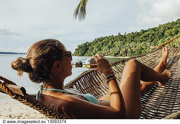 Junge Frau trinkt Alkohol  während sie sich in Hängematte am Strand ausruht