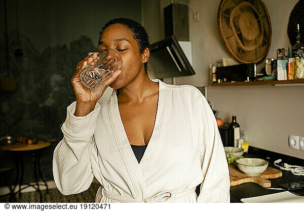 Junge Frau trinkt Wasser  während sie in der Küche zu Hause steht