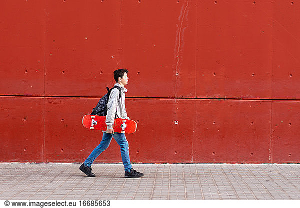 Junge hält ein rotes Skateboard  während er gegen eine rote Wand läuft