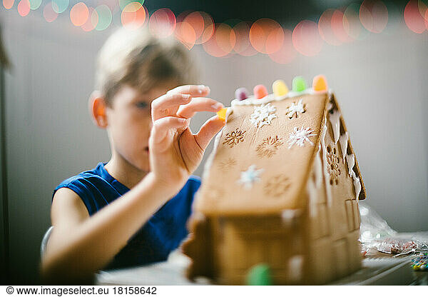 Junge legt Süßigkeiten auf Lebkuchenhaus mit Weihnachtsbeleuchtung