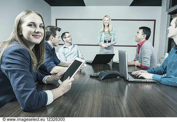 Junge Millennial-Geschäftsfrau  die in die Kamera schaut  während eine andere Frau eine Präsentation in einem Konferenzraum in einem modernen Geschäftshaus hält; Sherwood Park  Alberta  Kanada