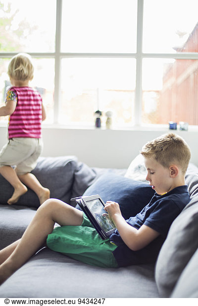 Junge mit digitalem Tablett auf dem Sofa  Schwester spielt im Hintergrund
