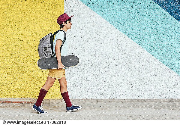 Junge mit Rucksack und Skateboard geht an einer bunten Wand vorbei