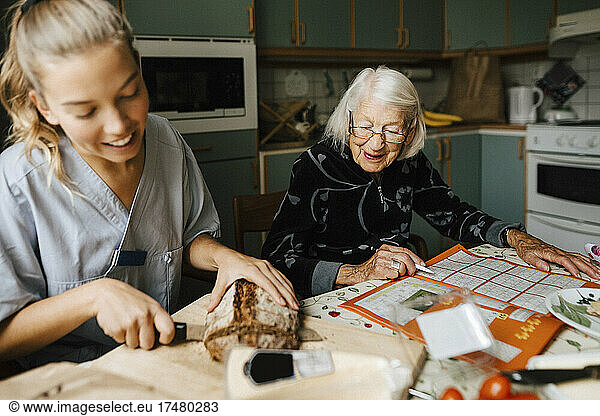 Junge Mitarbeiterin im Gesundheitswesen schneidet einen Brotlaib  während eine ältere Frau zu Hause ein Sudoku löst