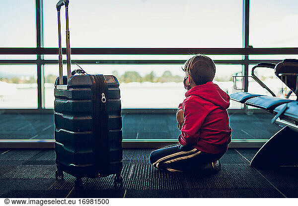 Junge neben einem Koffer  der am Flughafen aus dem Fenster schaut.