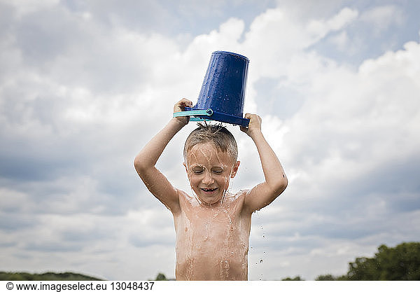 Junge ohne Hemd schüttet eimerweise Wasser über Kopf gegen bewölkten Himmel