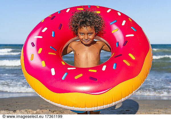 Junge ohne Hemd schaut durch aufblasbaren Donut am Strand