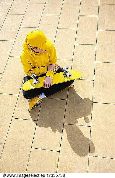 Junge schaut weg  während er mit dem Skateboard auf dem Fußweg sitzt  während eines sonnigen Tages