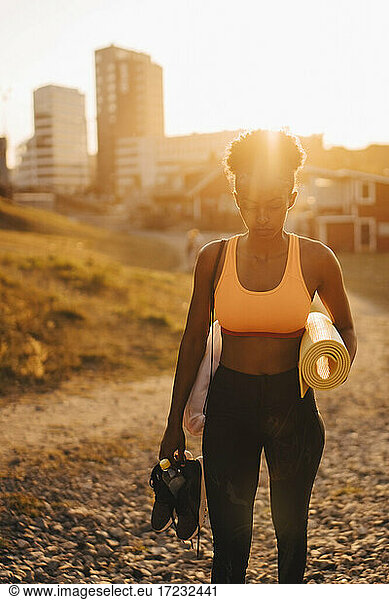 Junge Sportlerin mit Sportschuh und Trainingsmatte stehend an Land bei Sonnenuntergang