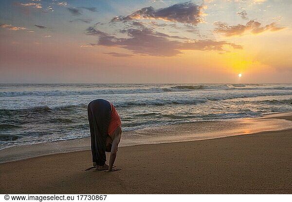 Junge sportliche fit Mann tun Yoga Sonnengruß Surya Namaskar Pose Hasta Uttanasana am tropischen Strand bei Sonnenuntergang. Mit Linsenreflexion