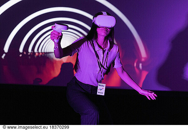 Junge Unternehmerin hält Joystick und schaut durch eine VR-Brille in einer beleuchteten Werkstatt