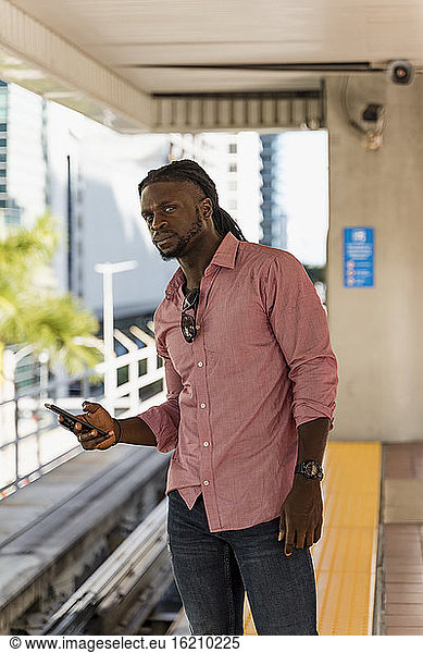 Junger Afroamerikaner  der sein Smartphone hält und wegschaut  während er am Bahnhof steht  Miami  Florida  USA