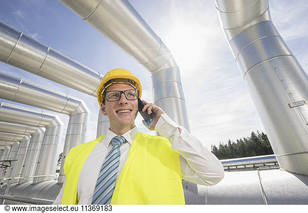 Junger Ingenieur  der in einem geothermischen Kraftwerk mit einem Mobiltelefon spricht