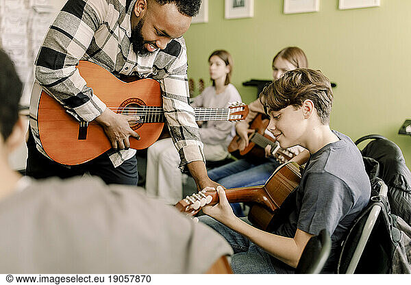 Junger männlicher Lehrer  der einem Teenager beim Gitarrenspiel im Musikunterricht an einer High School hilft