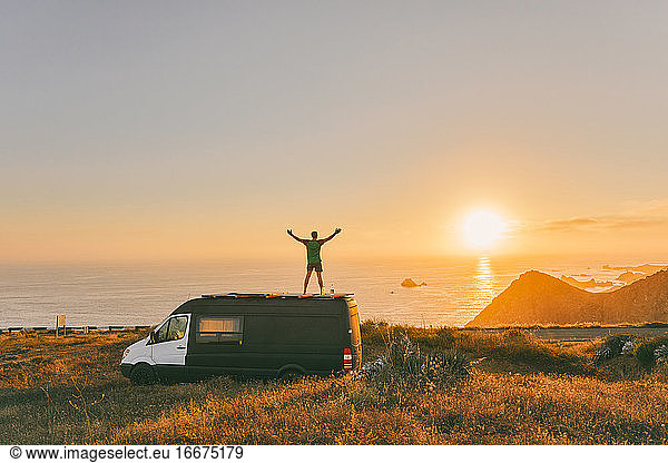 Junger Mann auf Sprinter-Van mit weit geöffneten Armen vor Sonnenuntergang auf Klippe in Baja  Mexiko.