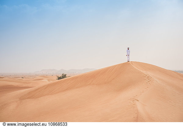 Junger Mann aus dem Nahen Osten in traditioneller Kleidung mit Blick auf Wüstendüne  Dubai  Vereinigte Arabische Emirate