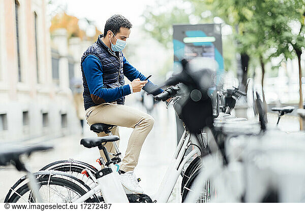 Junger Mann benutzt sein Smartphone zum Bezahlen eines Elektrofahrrads an einer Parkstation während einer Pandemie