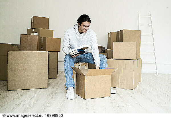 Junger Mann  der beim Auspacken eines Kartons in einem neuen Haus Bücher auspackt