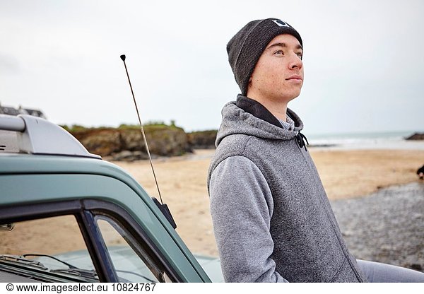 Junger Mann  der sich gegen das Auto lehnt und auf den Strand blickt  Constantine Bay  Cornwall  UK