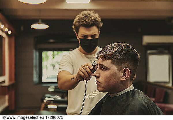 Junger Mann erhält einen Haarschnitt von einem männlichen Friseur in einem Salon während des COVID-19-Ausbruchs