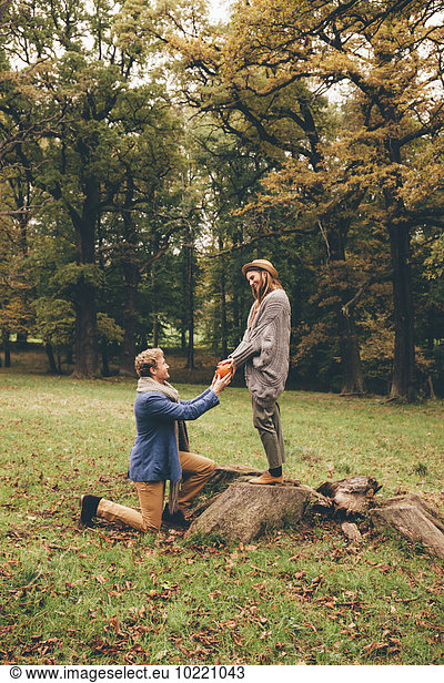 Junger Mann kniet nieder und macht seiner Freundin einen Antrag in einem herbstlichen Park.