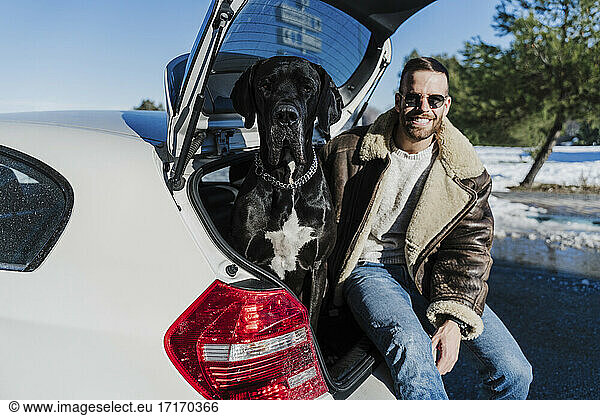 Junger Mann mit Hund sitzt im Kofferraum eines Autos an einem sonnigen Tag