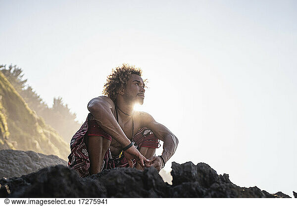 Junger Mann schaut weg  während er auf einem Felsen am Strand gegen den Himmel hockt