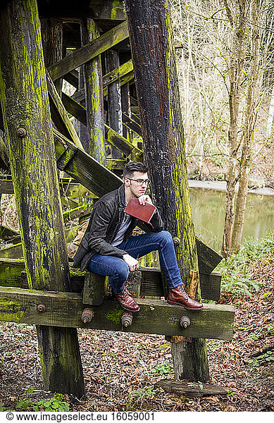 Junger Mann sitzt auf einer moosbewachsenen Gerüstbrücke neben einem Teich mit einem Buch in ruhiger Lage; Bothell  Washington  Vereinigte Staaten von Amerika