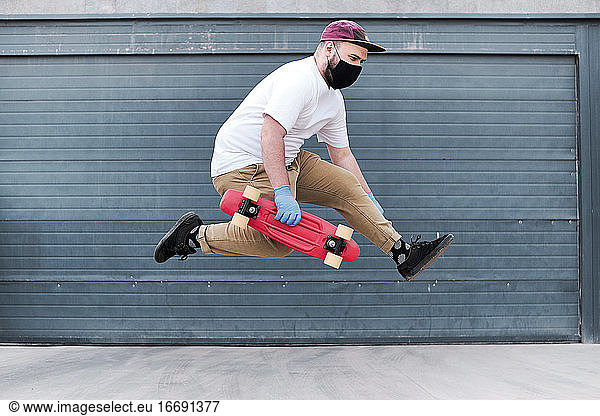 Junger Mann springt gegen eine schlichte blaue Garage und hält sein rotes Skateboard.